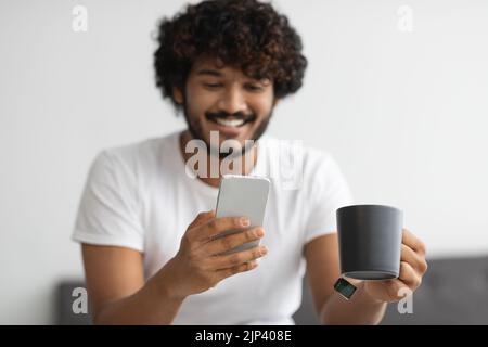 Lächelnder indischer Mann trinkt Kaffee und benutzt Smartphone Stockfoto