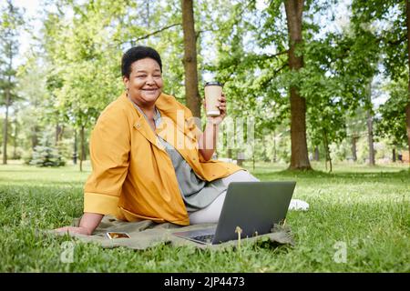 In voller Länge Porträt einer glücklichen reifen Frau mit Laptop im Freien, während Sie Picknick mit Kaffee auf grünem Gras genießen, Platz kopieren Stockfoto