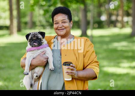 Waist-up-Porträt einer reifen schwarzen Frau mit niedlichem Mops im Freien und lächelnd vor der Kamera, während Sie gemeinsam im Park spazieren gehen Stockfoto
