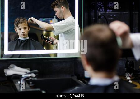 Junger männlicher Barbier, der im Friseurladen die Haare von Teenagern schneidet. Stockfoto