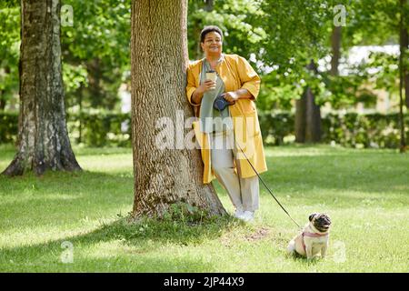 In voller Länge Porträt einer älteren schwarzen Frau, die sich im Park auf einen Baum lehnt, während sie mit einem niedlichen Hund spazieren geht und Kaffee trinkt, Platz zum Kopieren Stockfoto