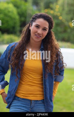 Porträt einer glücklichen Birazialfrau mit langen lockigen Haaren, die im Garten vor der Kamera lächelt Stockfoto