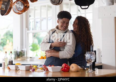 Glückliches biracial Paar, das Essen zubereitet, lächelt und umarmt in der Küche Stockfoto