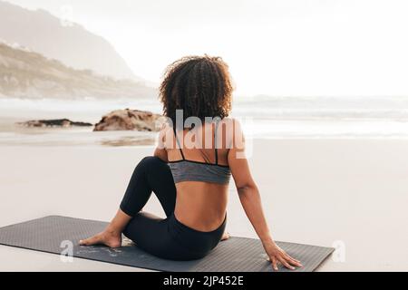Frau, die bei Sonnenuntergang am Strand auf einer Matte sitzt. Rückansicht einer Frau, die sich nach den Yoga-Übungen entspannt. Stockfoto