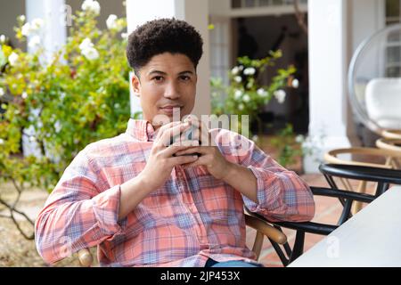 Glücklicher Birazialmann, der auf der Gartenterrasse sitzt und vor dem Haus Kaffee trinkt Stockfoto