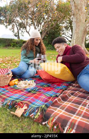 Vertikales Bild eines glücklichen, vielfältigen Paares, das im Herbstpark picknickt Stockfoto