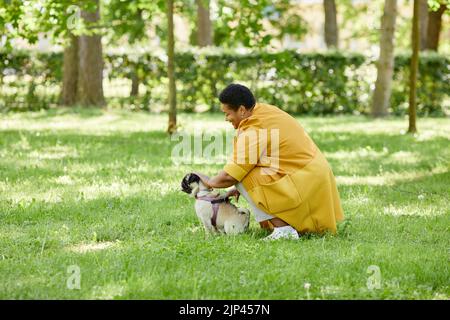 Seitenansicht Porträt von reifen schwarzen Frau Ausbildung niedlichen Hund im grünen Park und lächelnd, Copy Space Stockfoto