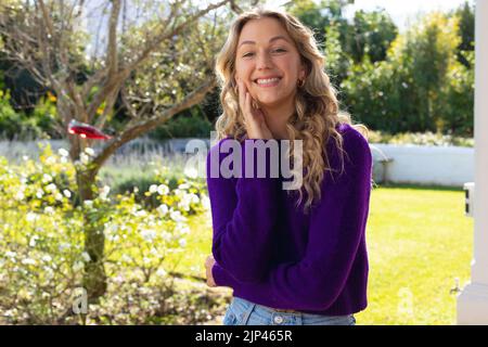 Porträt einer lächelnden kaukasischen Frau mit langen, lockigen Haaren im sonnigen Garten Stockfoto