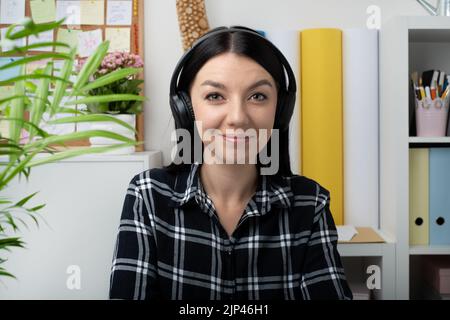 Eine fröhlich lächelnde junge Frau, die kabellose Kopfhörer trägt, schaut auf die Kamera. Online-Arbeiten am Arbeitsplatz vom Home Office aus. Stockfoto
