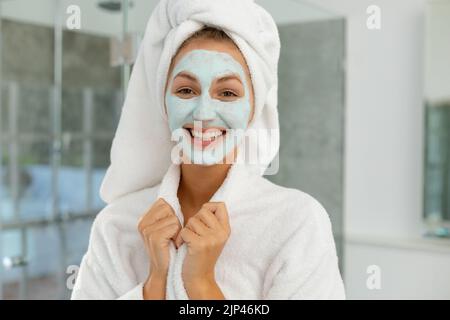 Porträt einer glücklichen kaukasischen Frau, die Robe trägt und im Badezimmer eine Schönheitsmaske aufträgt Stockfoto