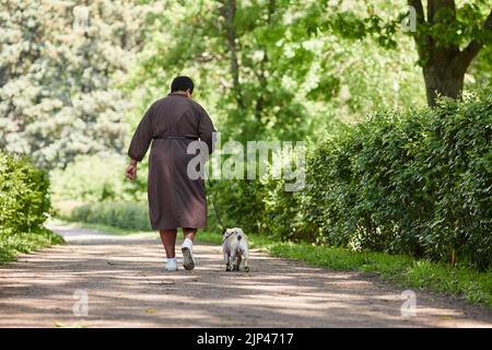Rückansicht Porträt einer schwarzen reifen Frau, die gemeinsam im Park auf dem Straßenweg den Hund läuft, Raum kopieren Stockfoto