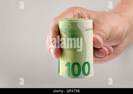 Die Hand hält eine Rolle von 100-Euro-Banknoten. Euro-Banknoten gerollt in der Hand eines weißen Mannes auf einem grauen Hintergrund. Das Konzept der finanziellen Unterstützung Stockfoto