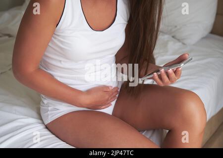 Junge Frau, die zu Hause an Menstruationskrämpfen leidet, sitzt auf dem Bett und blickt auf das Smartphone. Stockfoto