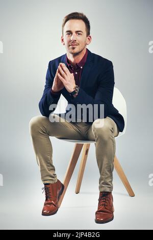 Sitzen im Sitz des Erfolgs. Studioporträt eines selbstbewussten jungen Geschäftsmannes, der auf einem Stuhl vor grauem Hintergrund sitzt. Stockfoto