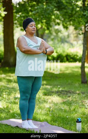 Vertikale Ganzkörperportrait der reifen schwarzen Frau, die Yoga im Freien im grünen Park genießt und mit geschlossenen Augen bei Atemübungen steht Stockfoto