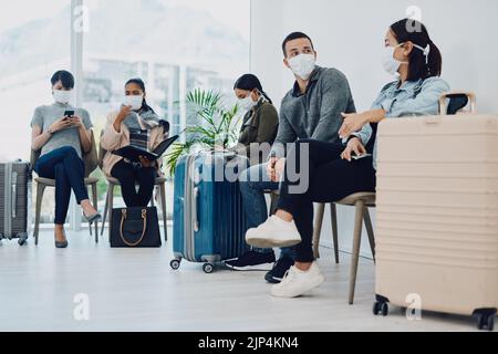 Eine Gruppe von Menschen, die während der Wartezeit in einer Flughafenlounge mit Schutzmasken unterwegs sind. Touristen, die bei einem öffentlichen Reiseverkehr in einer Schlange sitzen Stockfoto