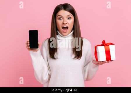 Aufgeregt überrascht Frau mit eingepackten Geschenk-Box und Handy mit leerem Bildschirm für die Förderung, trägt weißen lässigen Stil Pullover. Innenaufnahme des Studios isoliert auf rosa Hintergrund. Stockfoto