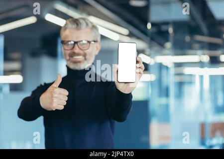 Erfolgreicher grauhaariger Senior Geschäftsmann zeigt Telefonbildschirm auf weißem Hintergrund, Mann im modernen Büro hält Daumen hoch empfehlen, reifer Investor Blick auf die Kamera Stockfoto