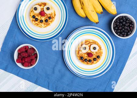 Amerikanische Pfannkuchen mit Himbeeren, Blaubeeren und Banane, dekoriert wie ein Lächeln und fröhliche Gesichter. Essen für Kinder, verspielt und kreativ. Draufsicht. Stockfoto