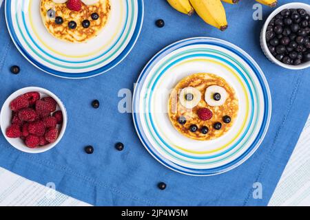 Amerikanische Pfannkuchen mit Himbeeren, Blaubeeren und Banane, dekoriert wie ein Lächeln und fröhliche Gesichter. Essen für Kinder, verspielt und kreativ. Draufsicht. Stockfoto