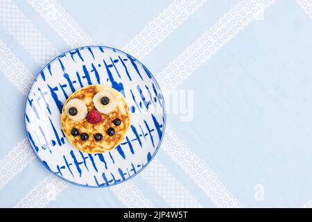 Amerikanische Pfannkuchen mit Himbeeren, Blaubeeren und Banane, dekoriert wie ein Lächeln und fröhliche Gesichter. Essen für Kinder, verspielt und kreativ. Draufsicht. Stockfoto