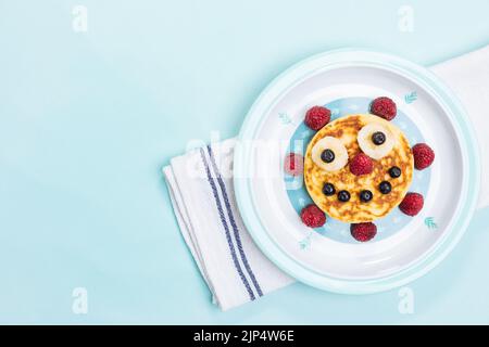 Amerikanische Pfannkuchen mit Himbeeren, Blaubeeren und Banane, dekoriert wie ein Lächeln und fröhliche Gesichter. Essen für Kinder, verspielt und kreativ. Draufsicht. Stockfoto