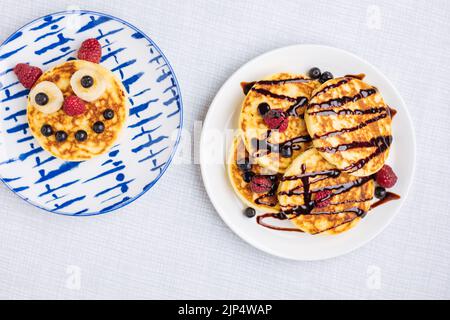 Amerikanische Pfannkuchen mit Himbeeren, Blaubeeren und Banane, dekoriert wie ein Lächeln und fröhliche Gesichter. Essen für Kinder, verspielt und kreativ. Draufsicht. Stockfoto