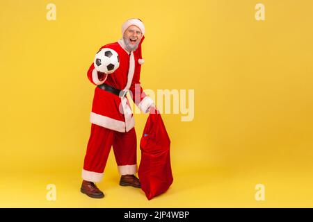 In voller Länge älterer Mann mit grauem Bart trägt weihnachtsmann Kostüm stehend mit Fußball und große rote Tasche mit Weihnachtsgeschenken. Innenaufnahme des Studios isoliert auf gelbem Hintergrund. Stockfoto