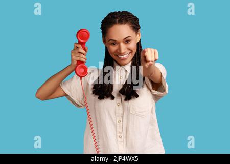 Schöne Frau mit schwarzen Dreadlocks, die Finger auf die Kamera zeigen, Hörer des roten Vintage-Festnetztelefons halten, Anrufe entgegennehmen, weißes Hemd tragen. Innenaufnahme des Studios isoliert auf blauem Hintergrund. Stockfoto