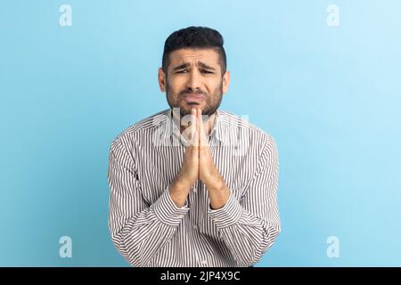 Das Porträt eines Geschäftsmannes hält die Handflächen zusammengedrückt, fleht den Engel an, hat unschuldige Ausdrücke, bittet um Entschuldigung, trägt ein gestreiftes Hemd. Innenaufnahme des Studios isoliert auf blauem Hintergrund. Stockfoto