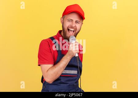 Porträt eines glücklichen, aufgeregten Arbeiters, der mit einem Mikrofon in den Händen steht und auf einer Firmenfeier singt und Spaß an der Arbeit mit Kollegen hat. Innenaufnahme des Studios isoliert auf gelbem Hintergrund. Stockfoto