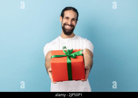 Porträt eines gutaussehenden entzückten Mannes mit Bart in weißem T-Shirt, Geschenkbox verpackt und lächelnd auf die Kamera, glücklicher Urlaub. Innenaufnahme des Studios isoliert auf blauem Hintergrund. Stockfoto