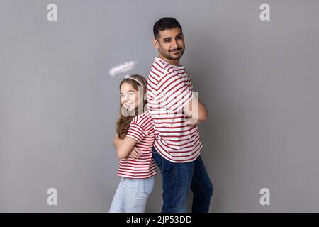 Porträt von lieblichen Vater und Tochter in gestreiften T-Shirts, Mann und Kind mit Halo über dem Kopf stehen Rücken an Rücken mit gefalteten Händen. Innenaufnahme des Studios isoliert auf grauem Hintergrund. Stockfoto