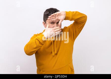 Fingerrahmen. Porträt eines Mannes, der durch den Handrahmen die Kamera anschaut, das Bild macht, das Auge fokussiert und einen Kapuzenpullover im urbanen Stil trägt. Innenaufnahme des Studios isoliert auf weißem Hintergrund. Stockfoto