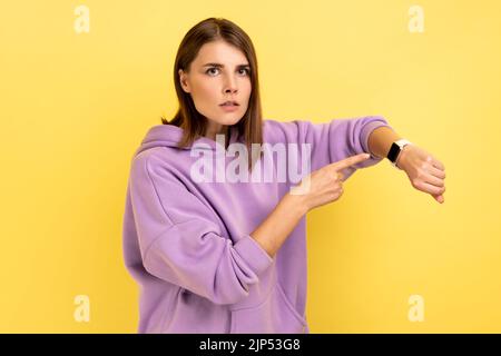 Porträt einer herrisch Frau mit dunklen Haaren, die auf die Armbanduhr zeigt und verärgert aussagt, zeigt die Uhr, die sich beeilen soll, trägt einen violetten Hoodie. Innenaufnahme des Studios isoliert auf gelbem Hintergrund. Stockfoto