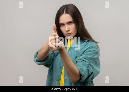 Ich werde Euch töten. Frau, die mit Fingerpistolen auf die Kamera zeigt, das Ziel anvisieren und mit Pistolenhandbewegung zu schießen droht, in einer Jacke im lässigen Stil. Innenaufnahme des Studios isoliert auf grauem Hintergrund. Stockfoto