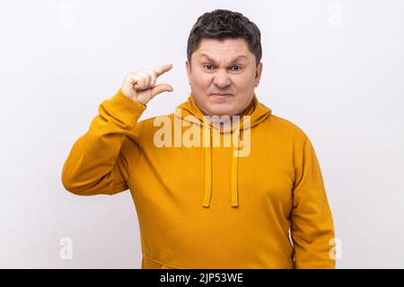 Porträt eines lustigen Mannes, der mit Fingern eine kleine Geste macht und sarkastisch lächelt, zeigt eine Mindestgröße, bettelt noch ein paar mehr, trägt einen Kapuzenpullover im urbanen Stil. Innenaufnahme des Studios isoliert auf weißem Hintergrund. Stockfoto