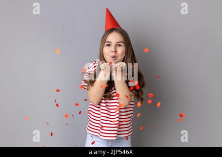 Portrait von glücklichen kleinen Mädchen trägt gestreiftes T-Shirt und Party-Kegel weht Herz geformt Konfetti, genießen Geburtstag, festliche Stimmung. Innenaufnahme des Studios isoliert auf grauem Hintergrund. Stockfoto