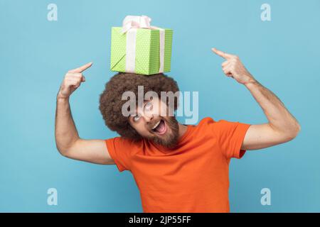 Porträt eines positiven, lustigen Mannes mit Afro-Frisur, das ein orangefarbenes T-Shirt trägt, das eine Geschenkbox auf dem Kopf hält und mit den Fingern zeigt und Aufregung ausdrückt. Innenaufnahme des Studios isoliert auf blauem Hintergrund. Stockfoto