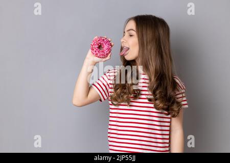 Porträt eines hungrigen kleinen Mädchens in gestreiftem T-Shirt, das Donut leckt und die Zunge zeigt, süßes zuckerhaltiges Süßwaren. Innenaufnahme des Studios isoliert auf grauem Hintergrund. Stockfoto