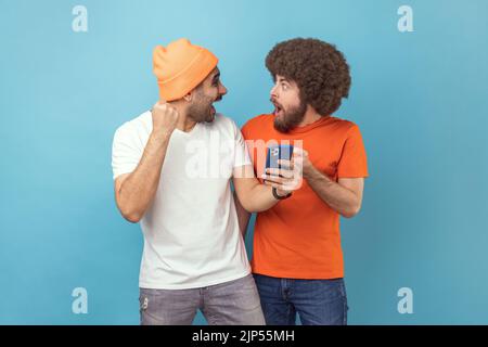 Porträt zweier aufgeregt staunender junger, erwachsener Hipster-Männer, die mit dem Handy stehen und Erfolge feiern, geballte Fäuste, die sich gegenseitig anguckend. Innenaufnahme des Studios isoliert auf blauem Hintergrund. Stockfoto