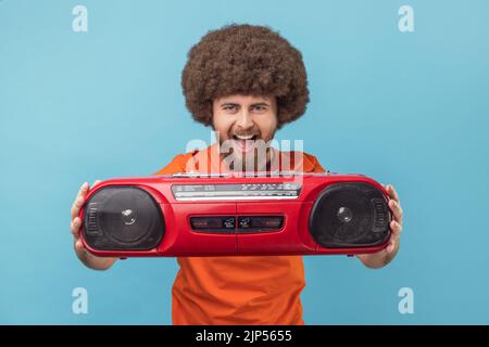 Porträt eines aufgeregten glücklichen Mannes mit Afro-Frisur, das ein orangefarbenes T-Shirt trägt und den roten Tonbandrekorder aushält und das Hören von Bändern genießt. Innenaufnahme des Studios isoliert auf blauem Hintergrund. Stockfoto