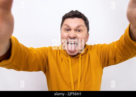 Porträt eines Mannes mittleren Alters, der ein Smartphone aus der Perspektive macht, die Zunge zeigt, kindhaftes Verhalten zeigt, POV, einen Hoodie im urbanen Stil trägt. Innenaufnahme des Studios isoliert auf weißem Hintergrund. Stockfoto