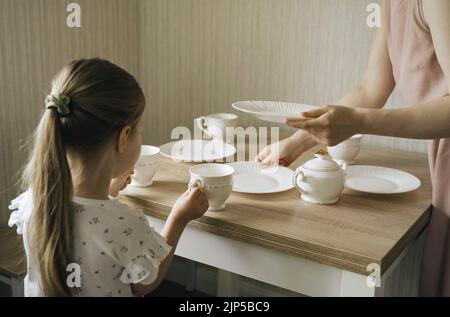 Mama und Tochter stellten Teller und Tassen auf den Tisch. Das Kind hilft seiner Mutter. Stockfoto