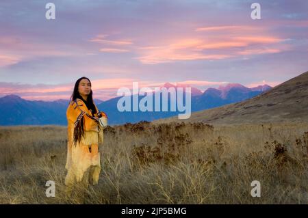Der Schwarzfuß-Mann, der in einem traditionellen, perlengeperlten Kriegshemd und Leggings gekleidet ist, steht auf einer Graswiese mit der Mission Mountain Range, Montana Stockfoto