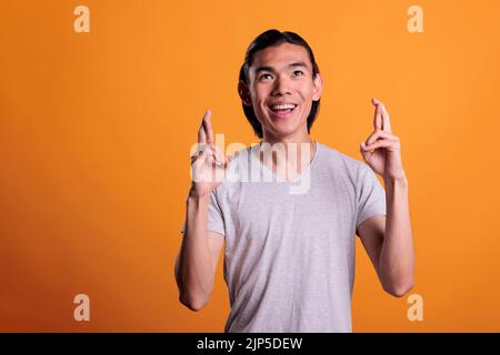 Junger asiatischer Mann, der die Finger kreuzt, um Glück betet und nach oben schaut. Lächelnder abergläubischer Teenager, der Glück hat, für Erfolg plädiert und mit hoffnungsvoller Mimik Wünsche macht Stockfoto