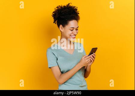 Zufriedene, fröhliche afroamerikanische junge Frau mit lockigen Haaren, die ihr Smartphone benutzt, auf den Bildschirm schaut, im Internet surft, mit Freunden chattet, auf isoliertem orangefarbenem Hintergrund steht und lächelt Stockfoto