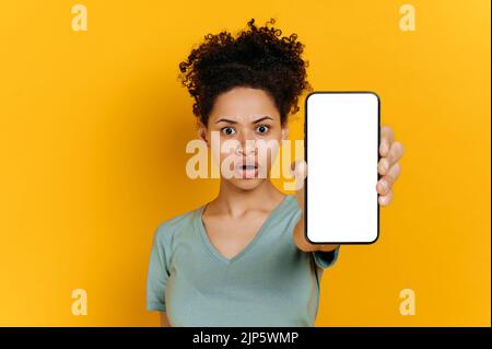 Emotional schockierte afroamerikanische junge Frau, in einem T-Shirt, hält ein Smartphone mit einem weißen Bildschirm in der Hand, steht auf einem isolierten orangefarbenen Hintergrund, blickt überrascht auf die Kamera. Modell Stockfoto