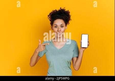 Schockiert verblüfft, die junge afroamerikanische lockige Frau, überrascht schaut auf die Kamera, hält das Handy mit leerem weißen Bildschirm und zeigt mit dem Finger darauf, steht auf isoliertem orangefarbenem Hintergrund Stockfoto