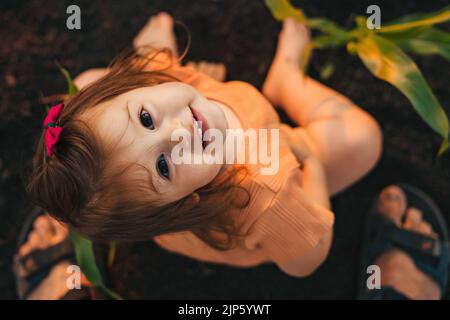 Baby-Mädchen, das aufschaut, während sie auf dem Boden im Kornfeld sitzt. Farmer Field. Glückliche Familie im Freien. Stockfoto